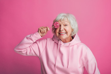 Positive, happy and cheerful old woman posing on camera alone. Stand and cover eye with lollipop. Wear stylish hoody. Isolated over pink background.
