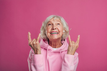 Emotional, cheerful and happy old woman in modern pink hoody posing on camera alone. Look up and...