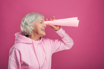 Positive cheerful old woman stand and look through rolled-up paper. Use it like spyglass or binoculars. Follow somebody. Wear modern hoody. Isolated over pink background. - obrazy, fototapety, plakaty