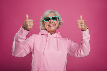 Positive old woman in pink hoody and black with green sunglasses posing on camera alone. Hold big thumbs up. Like symbol. Smile on her face with wrinkles. Isolated over pink background.