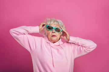 Confused old woman in modern pink hoody stand alone. Hold green and black sunglasses with hands....