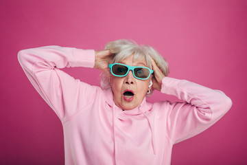 Terrified and scared old woman hold hands in hair and look straight through sunglasses. Wear pink...