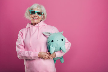 Funny, playful old woman in stylish pink sweater and green toy posing on camera. Wear sunglasses...