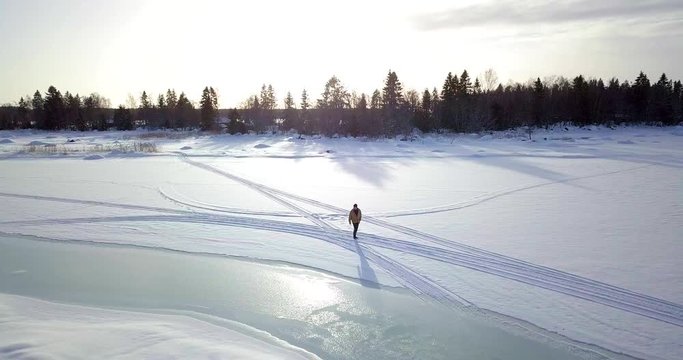 Aerial descending, tracking shot of lonely older man walking on icy frozen lake in winter, in Lapland, Finland