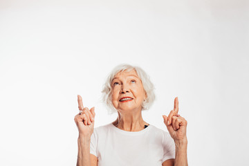 Cheerful, happy senior woman point up and look as well. Stand alone and pose on camera. Attractive female elder with grey hair. Isolated over white background.