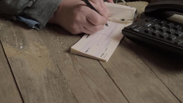 Hand of busy person writing a banking check to pay bills in office with phone
