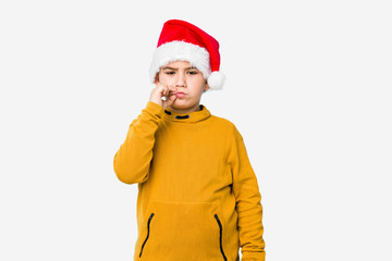 Little boy celebrating christmas day wearing a santa hat isolated with fingers on lips keeping a secret.