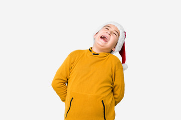 Little boy celebrating christmas day wearing a santa hat isolated relaxed and happy laughing, neck stretched showing teeth.