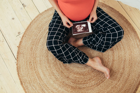 Cut view of barefeet young woman sitting on floor alone and hold ultrasound picture of her future baby. Single woman prepare to bacome mother. Maternity time soon.