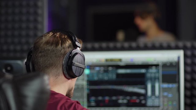 Rear view closeup of sound producer in earphones nodding head and giving thumbs up while listening to pop artist singing in booth in recording studio