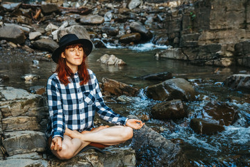 Beautiful girl with red hair in a hat and shirt meditating on rocks in a lotus position against a waterfall. Space for your text message or promotional content