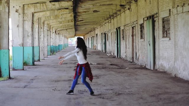 slim brunette with long loose hair and red checkered shirt on hips dances between abandoned house wall and columns