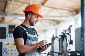 With notepad in hands. Industrial worker indoors in factory. Young technician with orange hard hat