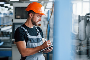 With notepad in hands. Industrial worker indoors in factory. Young technician with orange hard hat