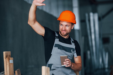 Hello gesture. With cup of drink. Lunch break. Cheerful industrial worker indoors in factory. Young...