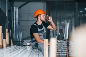 With cup of drink. Lunch break. Industrial worker indoors in factory. Young technician with orange...