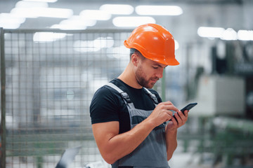 Smartphone in hands. Industrial worker indoors in factory. Young technician with orange hard hat