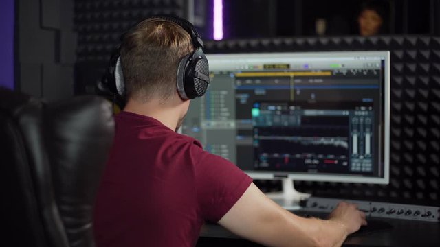 Back view zoom in of male sound producer in earphones using professional software on computer to record song in music production studio