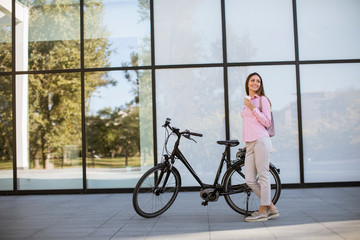 Beautiful young female cyclist drinks warm coffee from a cup by the electric bicycle in the urban...