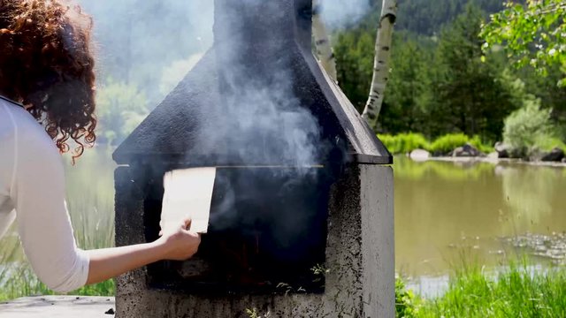 Girl blows coals and fans embers in the barbecue bbq brazier. Outdoors picnic concept.