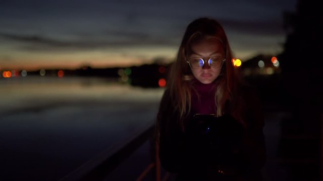 Sad young woman using her phone texting and standing on a pier - Vivid night on a river with bokeh light in the background - 1.4 aperture thin dop