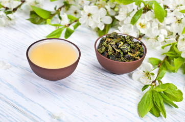 Green tea and dried leaves of tieguanyin tea in a ceramic cups with branches of blossoming apple tree on a white wooden background.