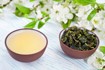 Green tea and dried leaves of green oolong tieguanyin tea in a ceramic cups with branches of blossoming apple tree on a white wooden background.