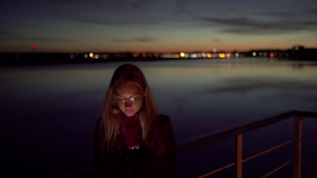 Sad young woman using her phone texting and standing on a pier - Vivid night on a river with bokeh light in the background - 1.4 aperture thin dop