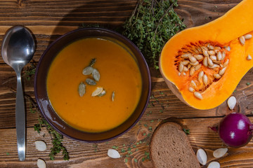 Top view on a plate of pumpkin soup on a wooden table with vegetables and a slice of bread.