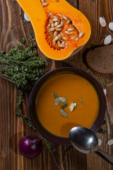 Top view on a plate with pumpkin soup on a wooden table with vegetables, thyme and seeds.