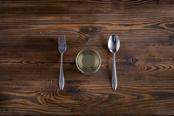 Closed metal tin can, fork and spoon on a brown wooden table background.