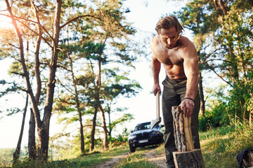 Woodsman with an axe cutting wood. Handsome shirtless man with muscular body type is in the forest at daytime