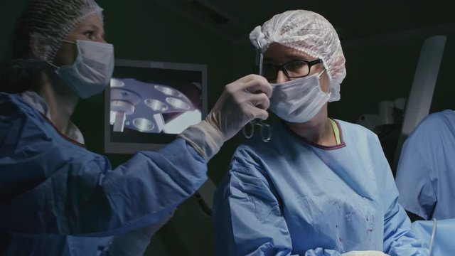 Pan with slow motion of nurse holding gauze and wiping sweat from forehead of female surgeon stitching patient