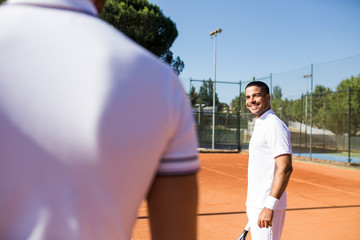 Man in sportswear smiling and looking at crop competitor while standing on tennis court