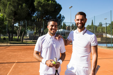 Two men in white sportswear holding rackets and smiling while standing on tennis court