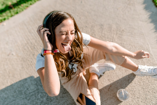 Young woman sitting in park using smartphone and listening to music with headphones and sticking out her tongue