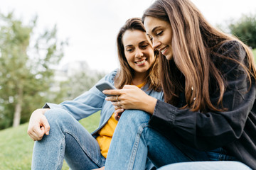 Two friends looking on the smartphone, sitting on a meadow of a park
