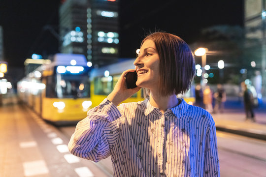 Young woman in the city at night talking on the phone, tram in the background, Berlin, Germany