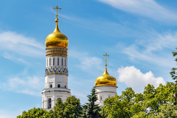 Fototapeta na wymiar Golden domes of Ivan the Great Bell-Tower and Uspenskaya belfry against blue sky with white clouds and green trees in sunlight. Moscow Kremlin architecture