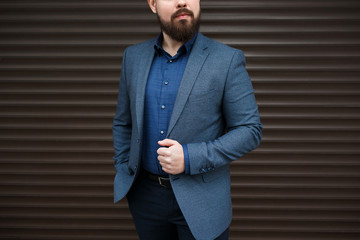 Stylish handsome brunette man with beard, wearing suit jacket and blue shirt, outdoors on the city street near brown roller door. Details of classic elegant formal men's outfit. Businessman.