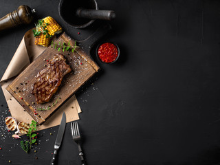 Fried steak with herbs and spices on wooden board, parchment, fork, knife, garlic, corn, sprouts, pepper, salt, sauce. Black background. Top view.
