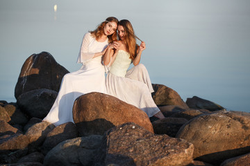 Two beautiful tender girls in long white wedding dresses are sitting on the stones by the sea in the evening.