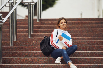 Positive female student sitting on stairs outdoors with documents at daytime