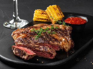 Sliced, fried, spiced juicy steaks with herbs on a dark board, grilled corn, red sauce and glass on a black background. Close-up shot. Side view.