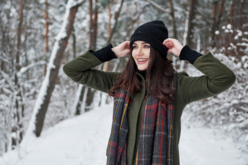 Fototapeta na wymiar Touches the black hat. Cheerful young girl in warm clothes have a walk in the winter forest at daytime