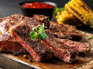 Sliced, fried, spiced steaks with herbs on wooden board, grilled corn, red sauce in small dark bowl...