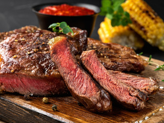 Sliced, fried, spiced steaks with herbs on wooden board, grilled corn, red sauce in small dark bowl...