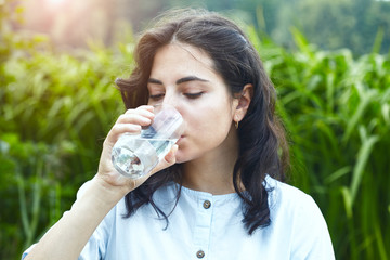 healthy beautiful young  brunette woman holding glass of clear pure water and drinking outside, morning light