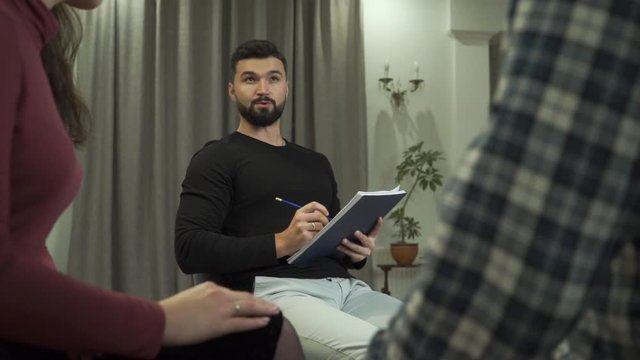 Highly professional Caucasian male psychologist looking through notes, and telling patient their results. Young couple or family taking each other hands at the foreground. Successful couples therapy.