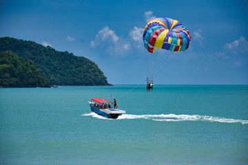 Parasailing on the waves of the azure Andaman sea under the blue sky near the shores of the sandy beautiful exotic and stunning Cenang beach in Langkawi island, - obrazy, fototapety, plakaty